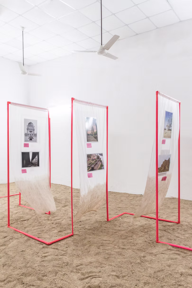 Three sheer fabric panels on hot-pink pipe frames displaying photographs, set on sand under ceiling fans, Hyderabad gallery