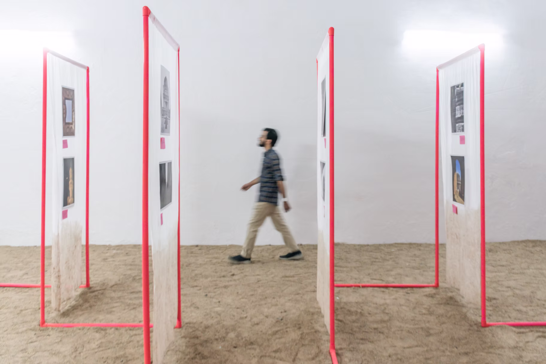 Red-framed doorway panels hung with photo prints over a sand floor, a visitor walking between them, Hyderabad installation