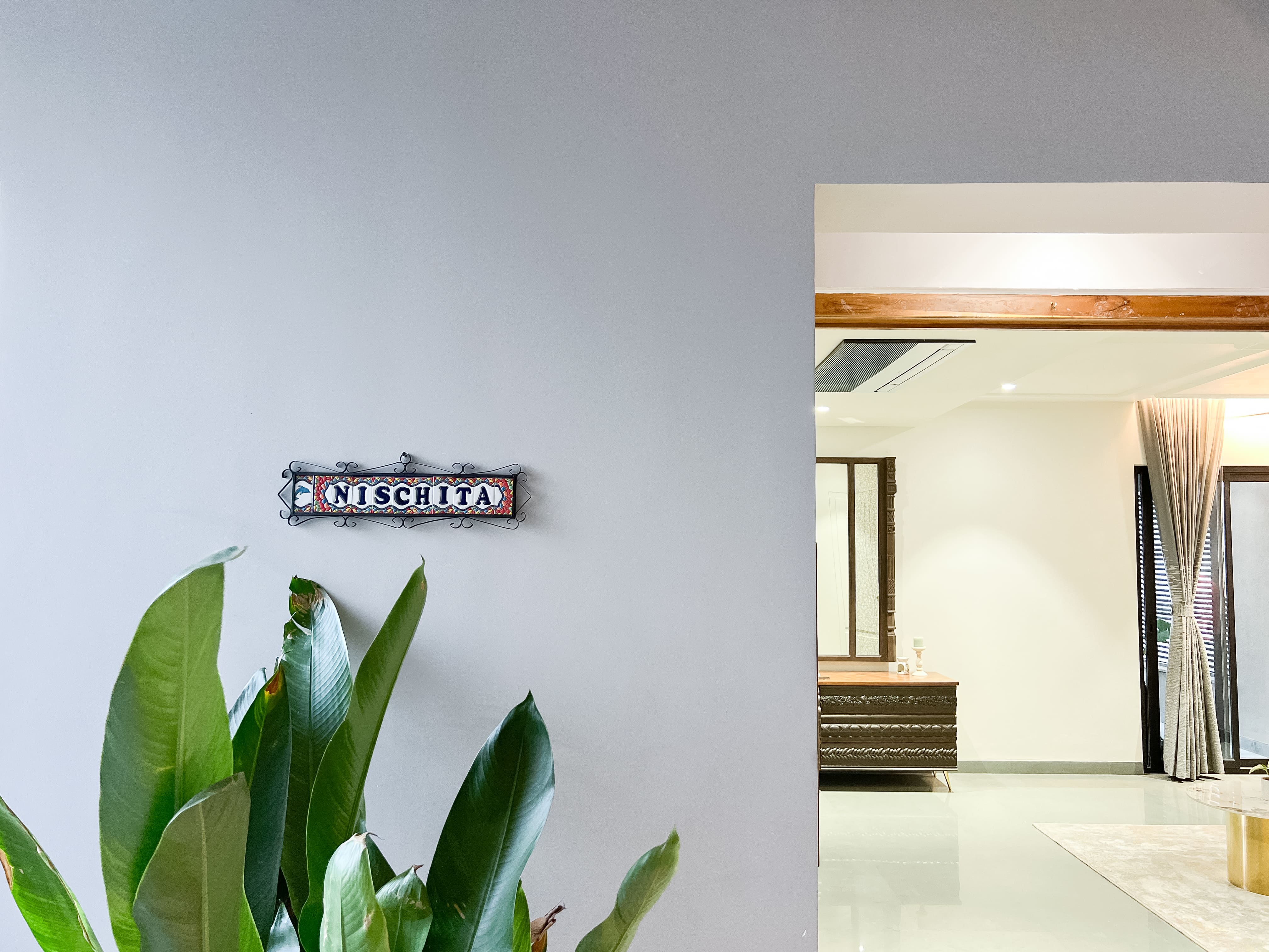 Carved metal nameplate reading NISCHITA mounted on a grey wall beside a leafy plant near a bright entryway, Hyderabad home