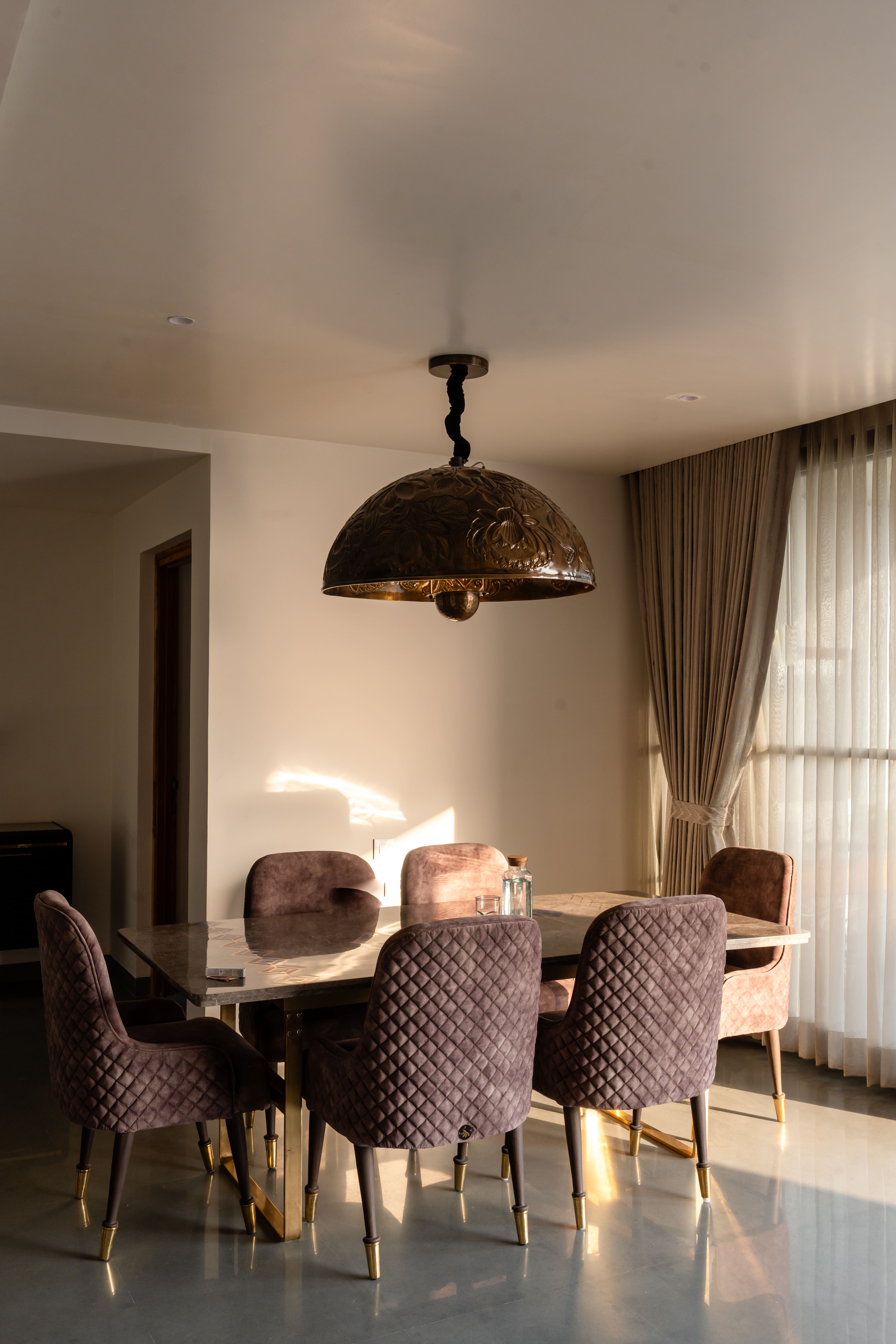 Dining area in Hyderabad with dusty-mauve velvet chairs around a marble table, copper dome pendant and pleated curtains