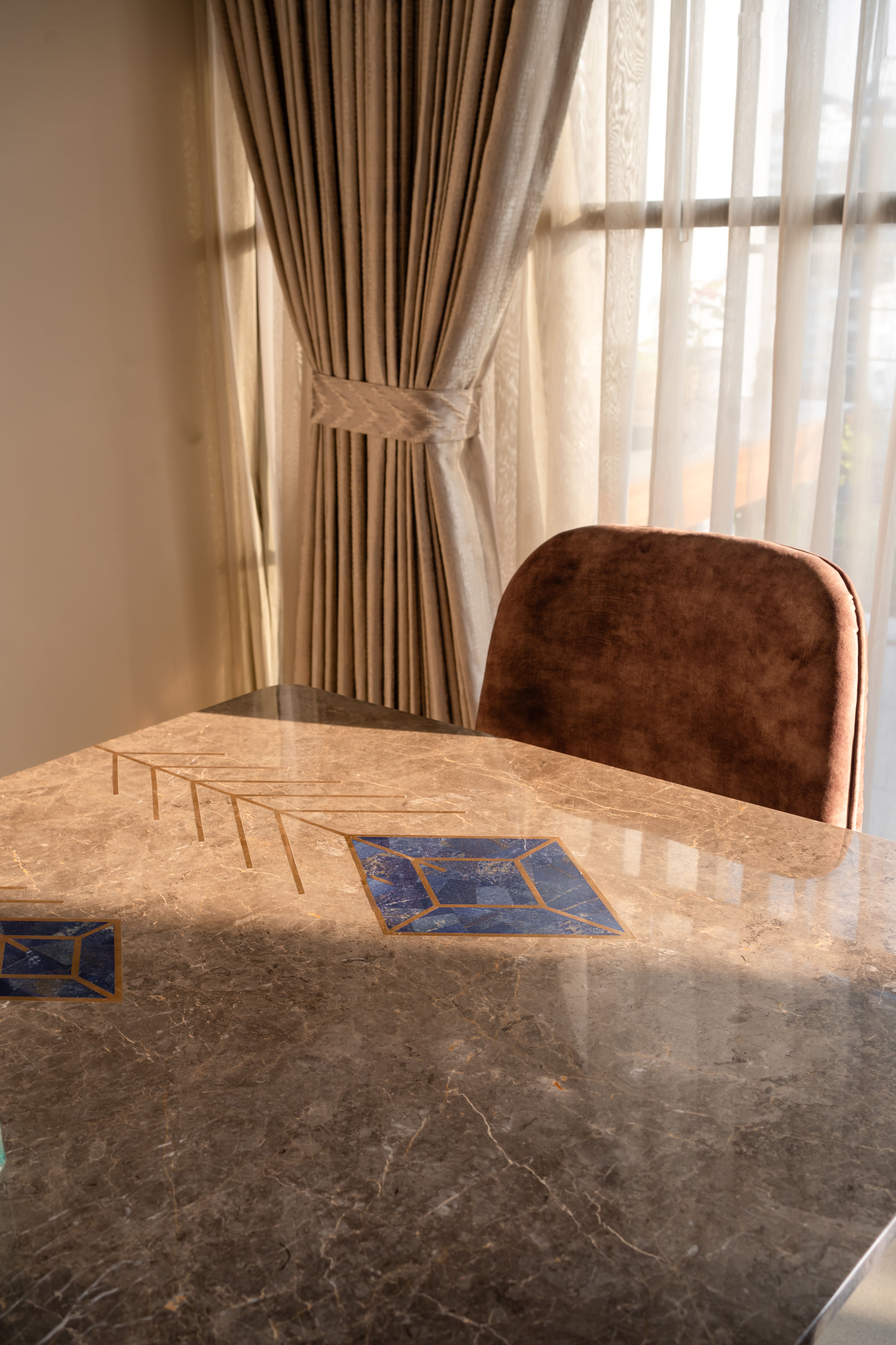 Close-up of a brown marble dining table with blue lapis-style inlay diamonds and brass trim beside draped curtains, Hyderabad