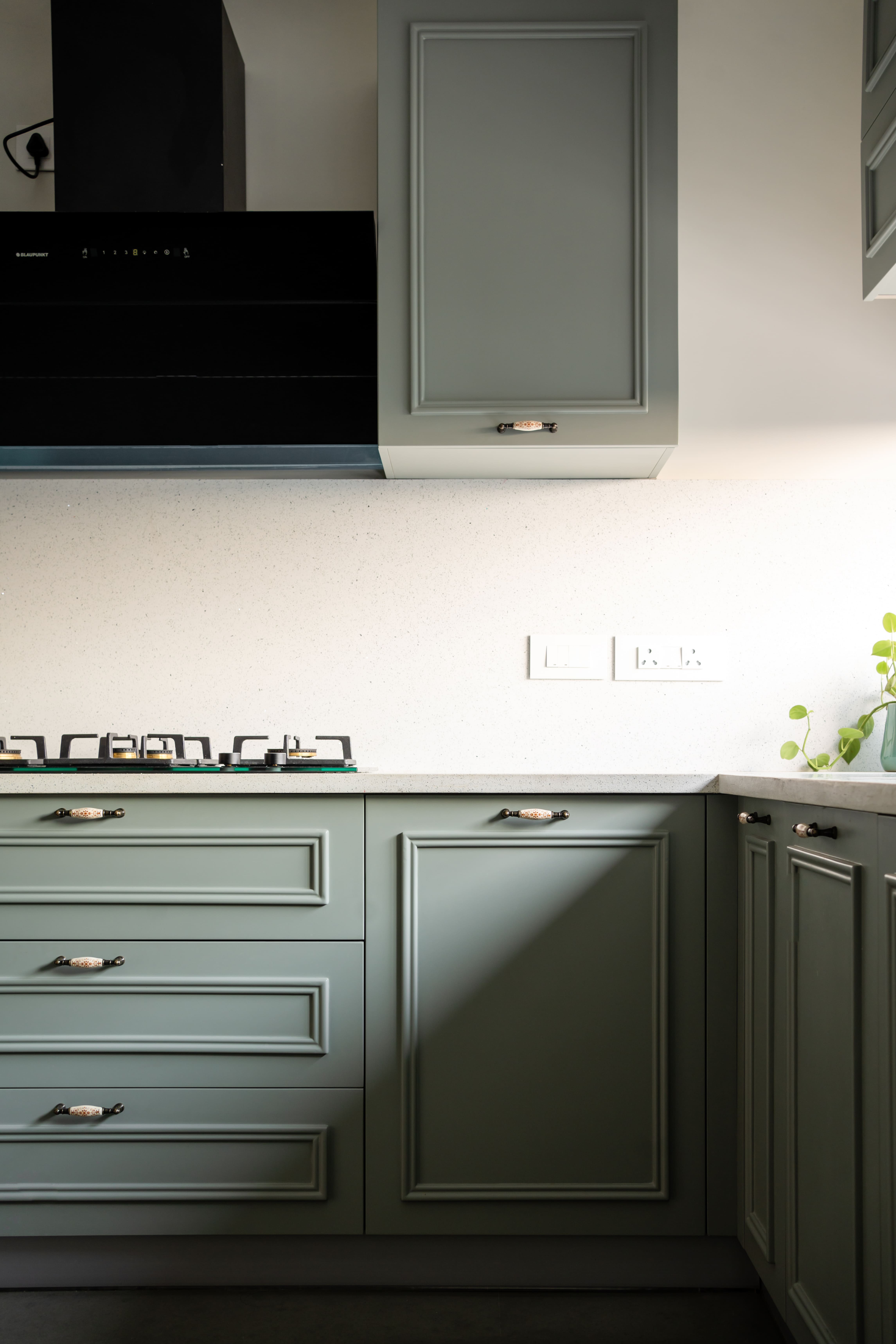 Sage-green kitchen detail, Hyderabad — paneled cabinets with cup pulls, black wall chimney and gas hob on a speckled quartz top