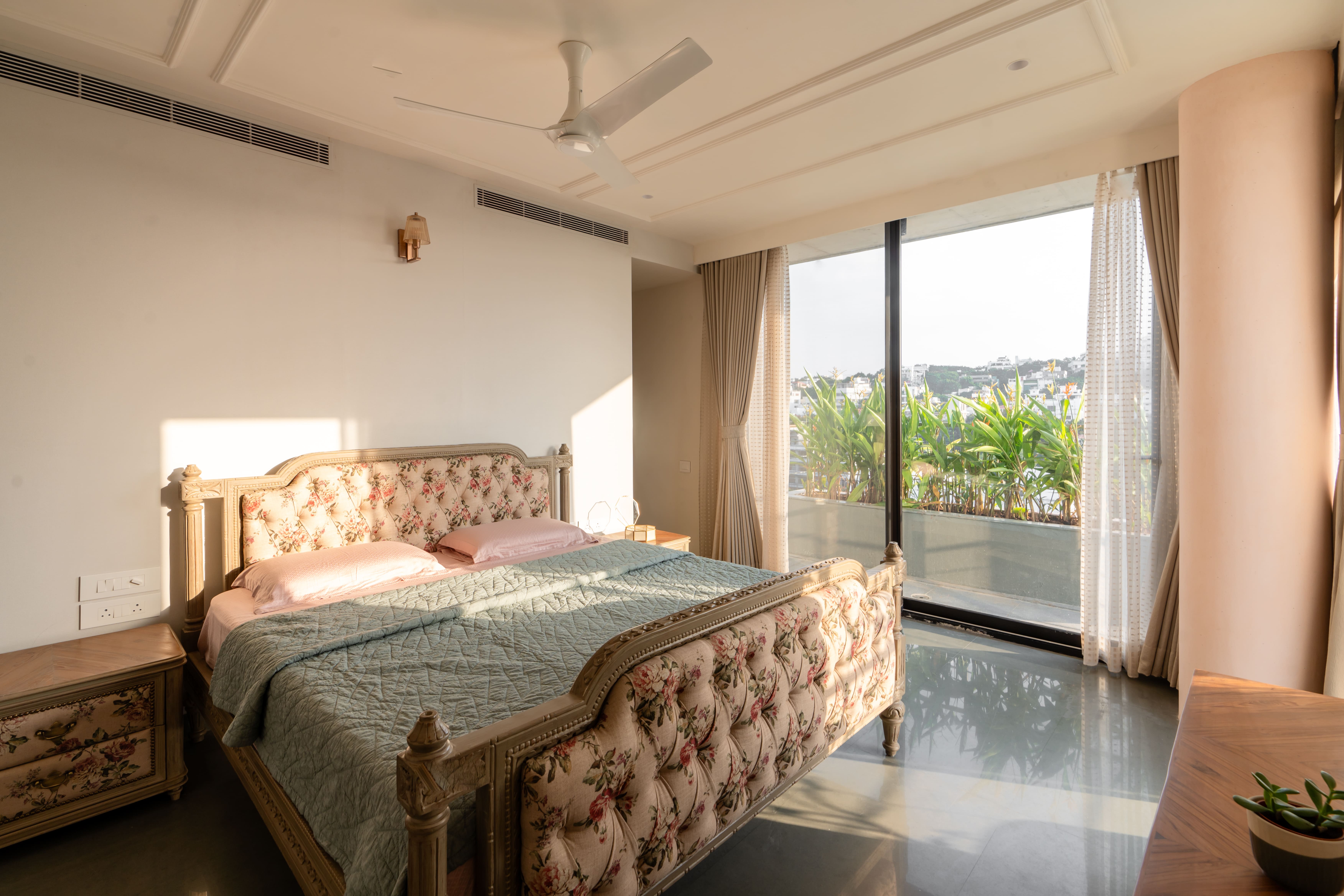 Master bedroom in Hyderabad with a floral tufted bed, pink pillows and a wide window overlooking the hills and greenery