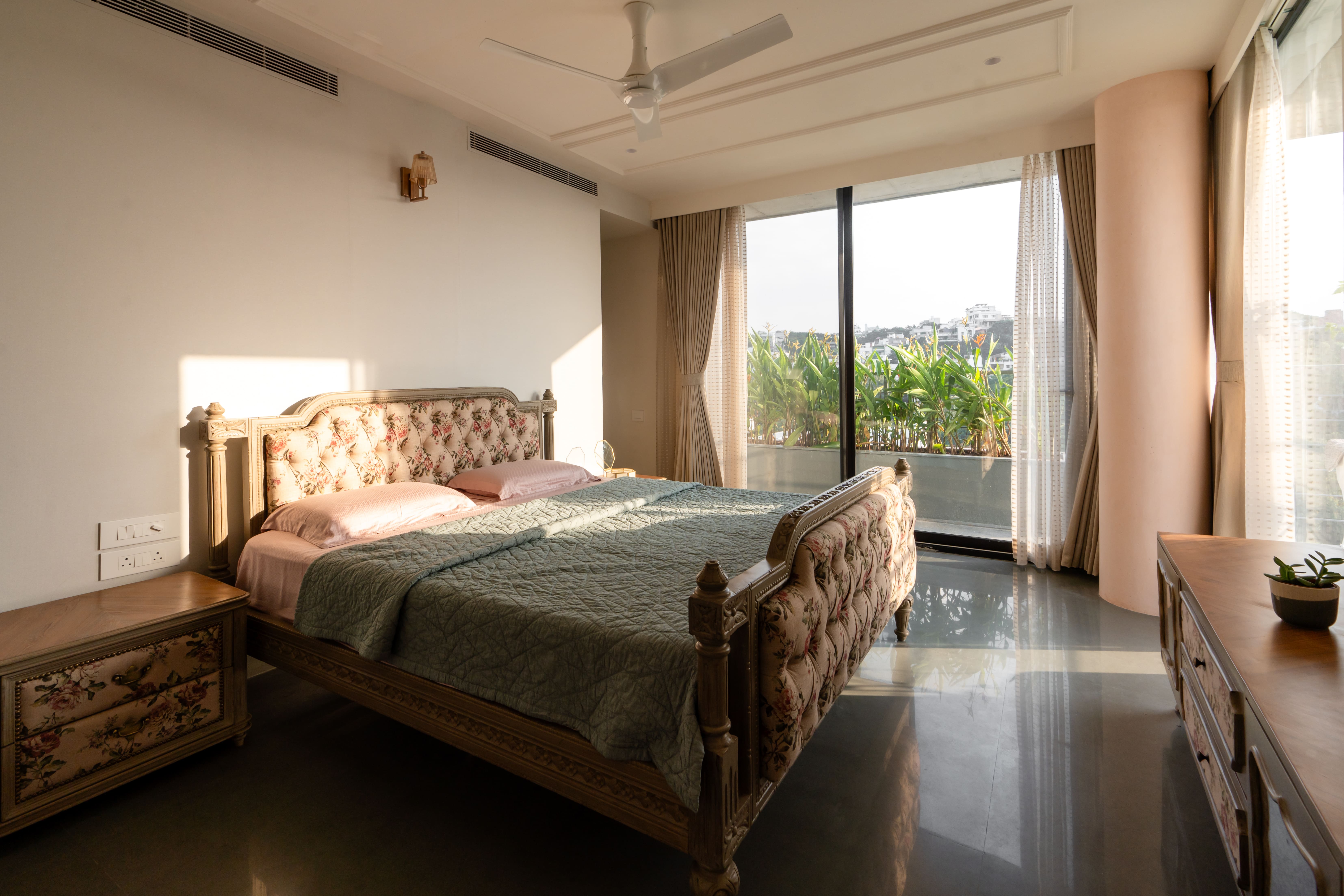 Spacious master bedroom, Hyderabad — tufted floral carved bed, green bedspread and full-height windows to a leafy balcony
