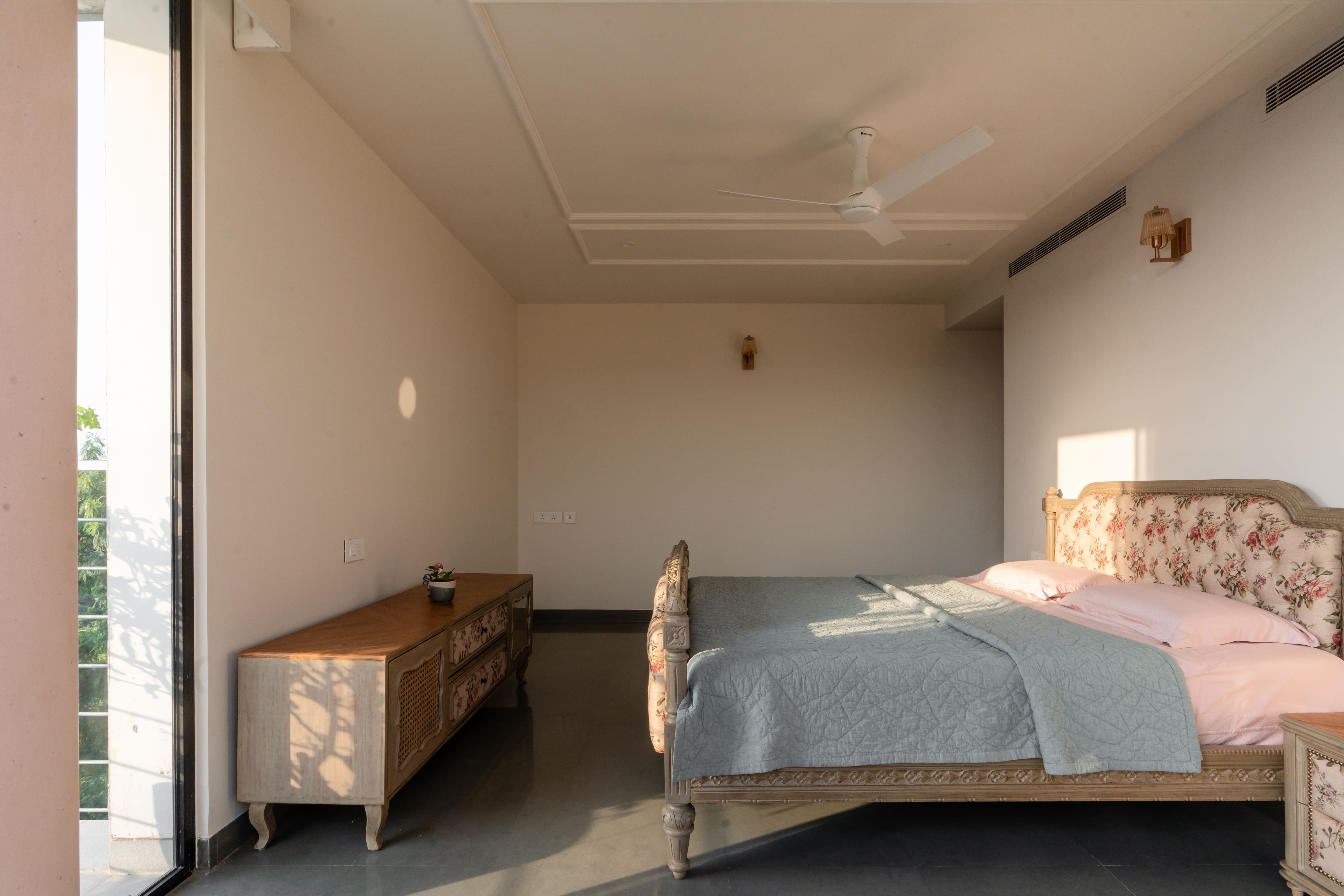 Master bedroom with floral upholstered bed, pink and grey bedding, carved wood console and balcony glass doors, Hyderabad