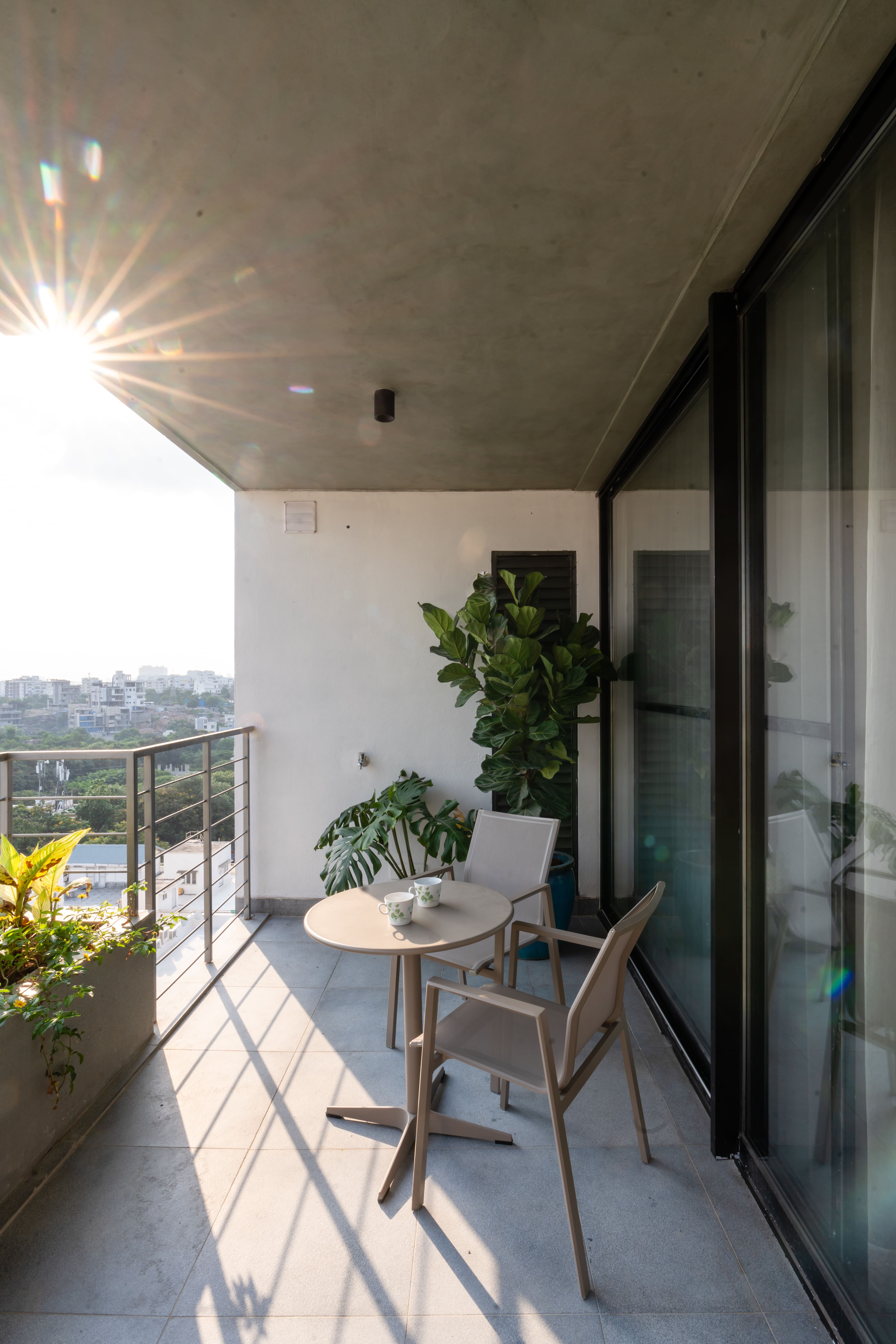 Sunlit balcony with two cane-style chairs, round table and tall indoor plant framed by glass sliding doors, Hyderabad
