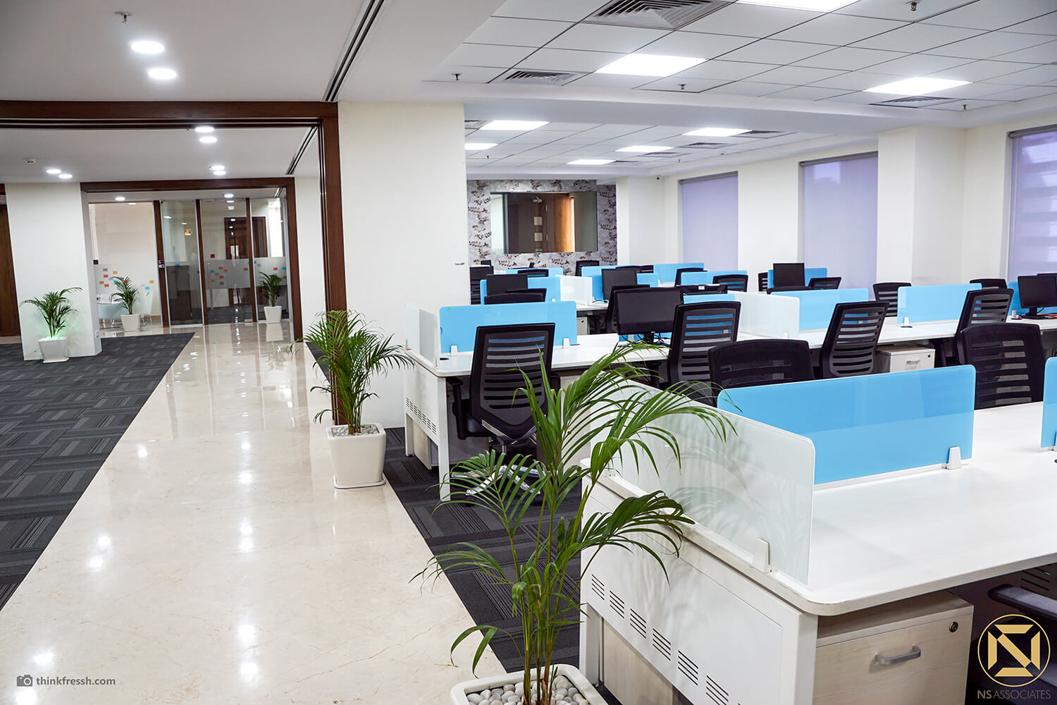 Open-plan office floor with blue-screened workstations, potted palms and polished tile aisle, Hyderabad workplace