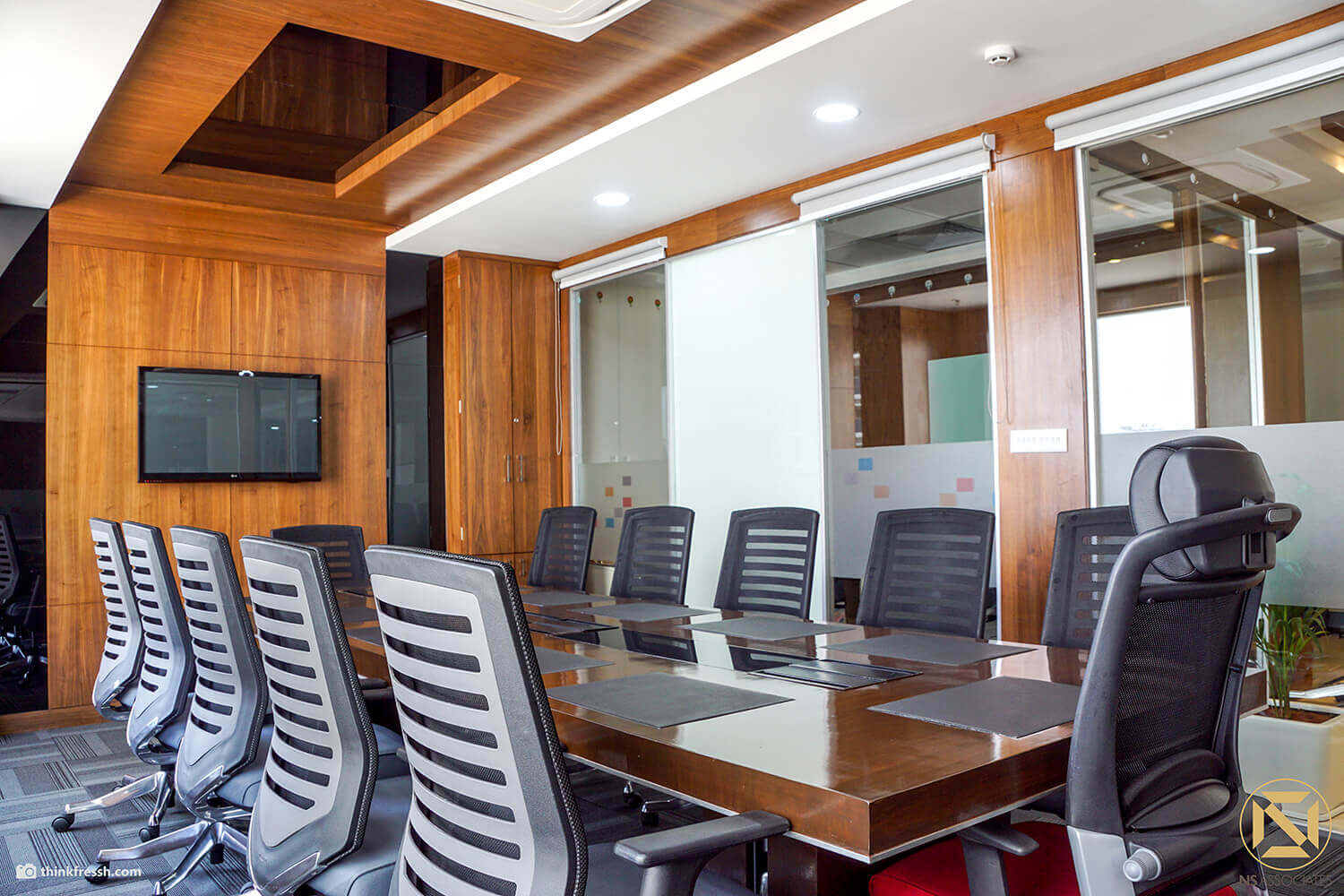 Office boardroom in Hyderabad with long wood-veneer table, black mesh chairs, wall-mounted TV and frosted glass partition
