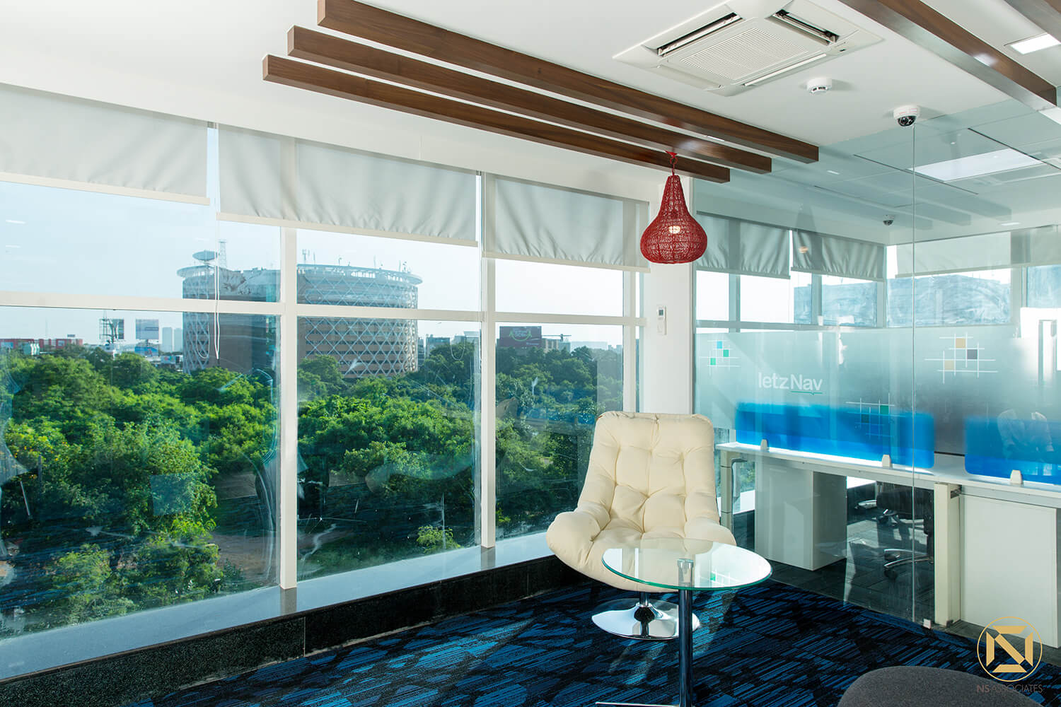 Breakout corner with cream tufted lounge chair, red pendant lamp and blue patterned carpet by panoramic windows, Hyderabad office