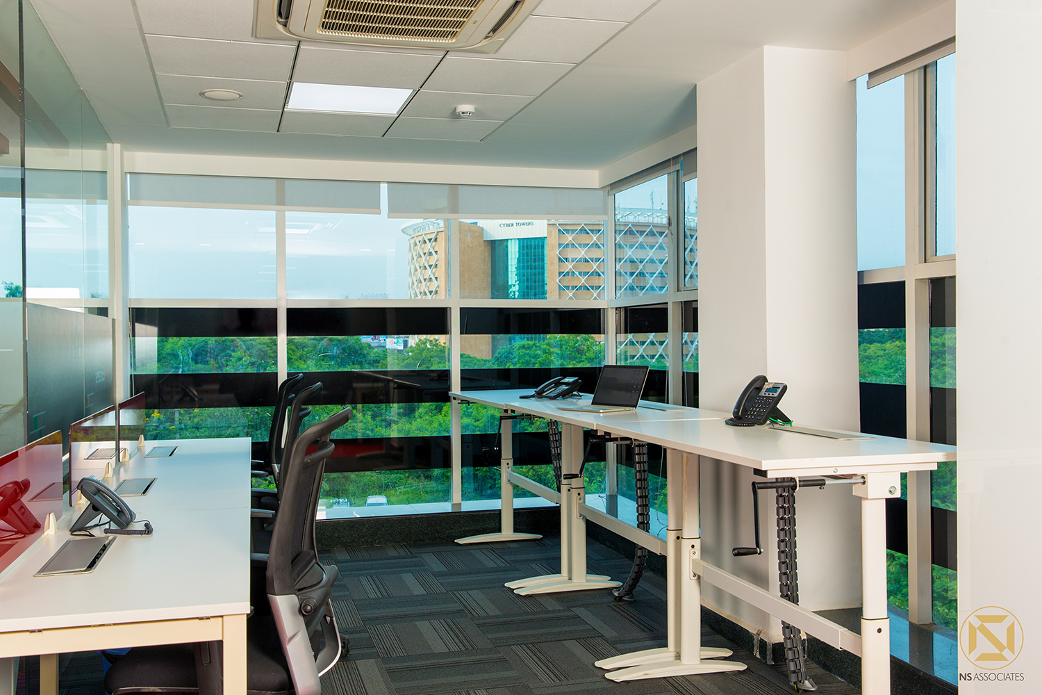 Open workstation bay with white height-adjustable desks and floor-to-ceiling tinted glazing overlooking the city, Hyderabad