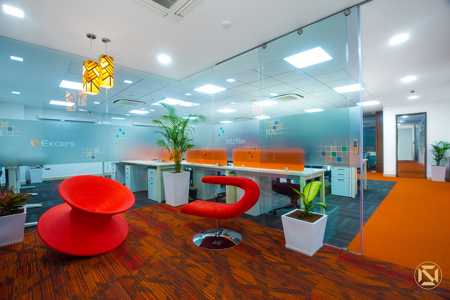 Office breakout zone with sculptural red chairs, yellow pendant lights and orange accents framed by glass partitions, Hyderabad