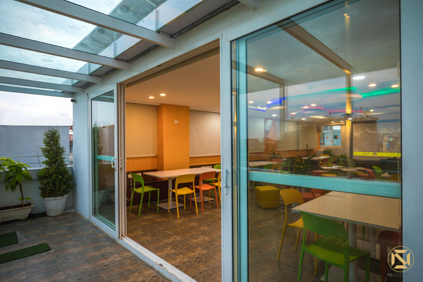 Rooftop terrace cafeteria with glass-roof canopy and colourful green-and-yellow chairs, viewed through sliding glass doors, Hyderabad office