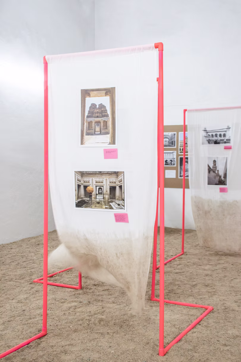Two pink-pipe panels with architecture prints and sheer fabric pooling onto a sand floor in a bright white gallery, Hyderabad