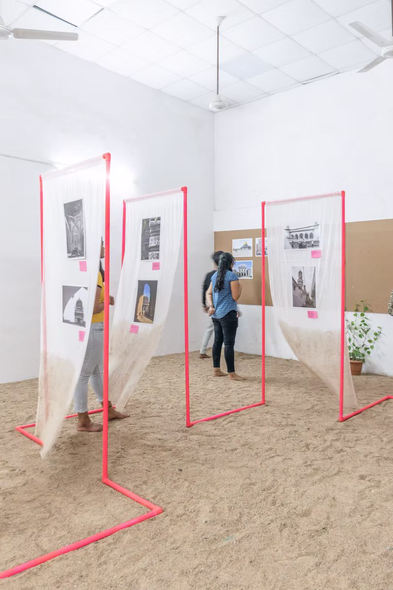 Wide gallery view of red doorway frames draped in gauze over sand, two visitors viewing the photo prints, Hyderabad