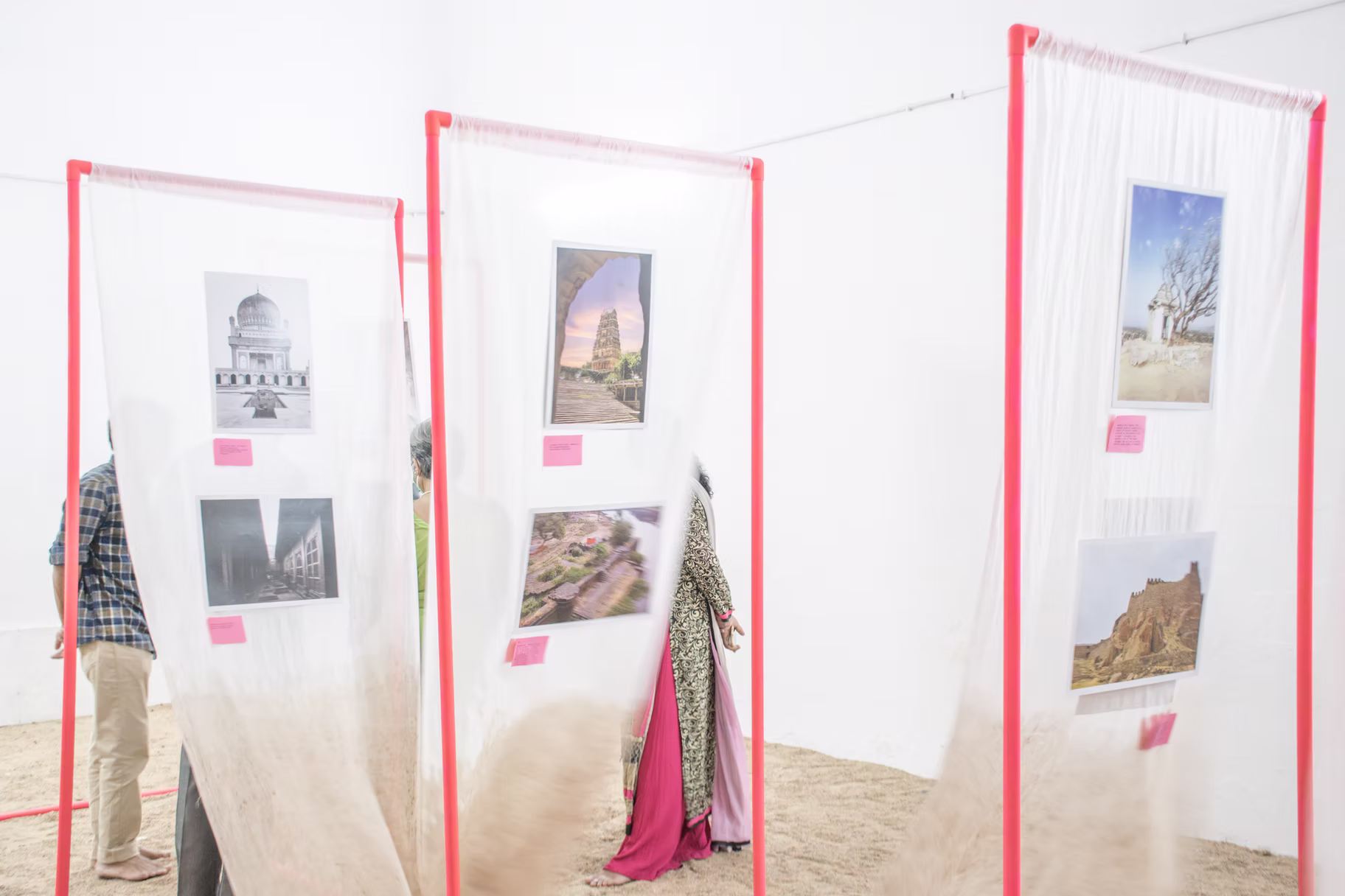 Sheer panels on pink frames displaying Hyderabad monument photos, fabric draping to a sand floor in a white gallery