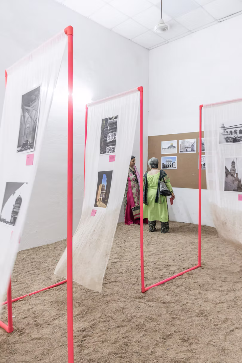 Gauze-draped red frames with monochrome photo prints, a visitor in green browsing a pinboard behind them on sand, Hyderabad