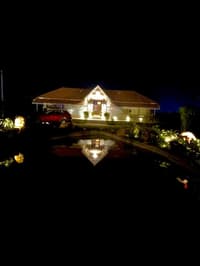Hyderabad stone farmhouse lit up at night, its warm facade lights mirrored in the still pond out front
