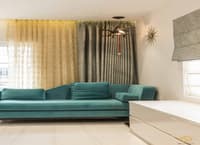 Living room corner in Hyderabad with low teal velvet sofa, layered sheer and grey drapes and starburst wall clock