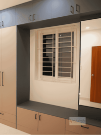 Window-seat storage bench flanked by grey and beige wardrobes with a mirror panel beside a grilled window, Hyderabad