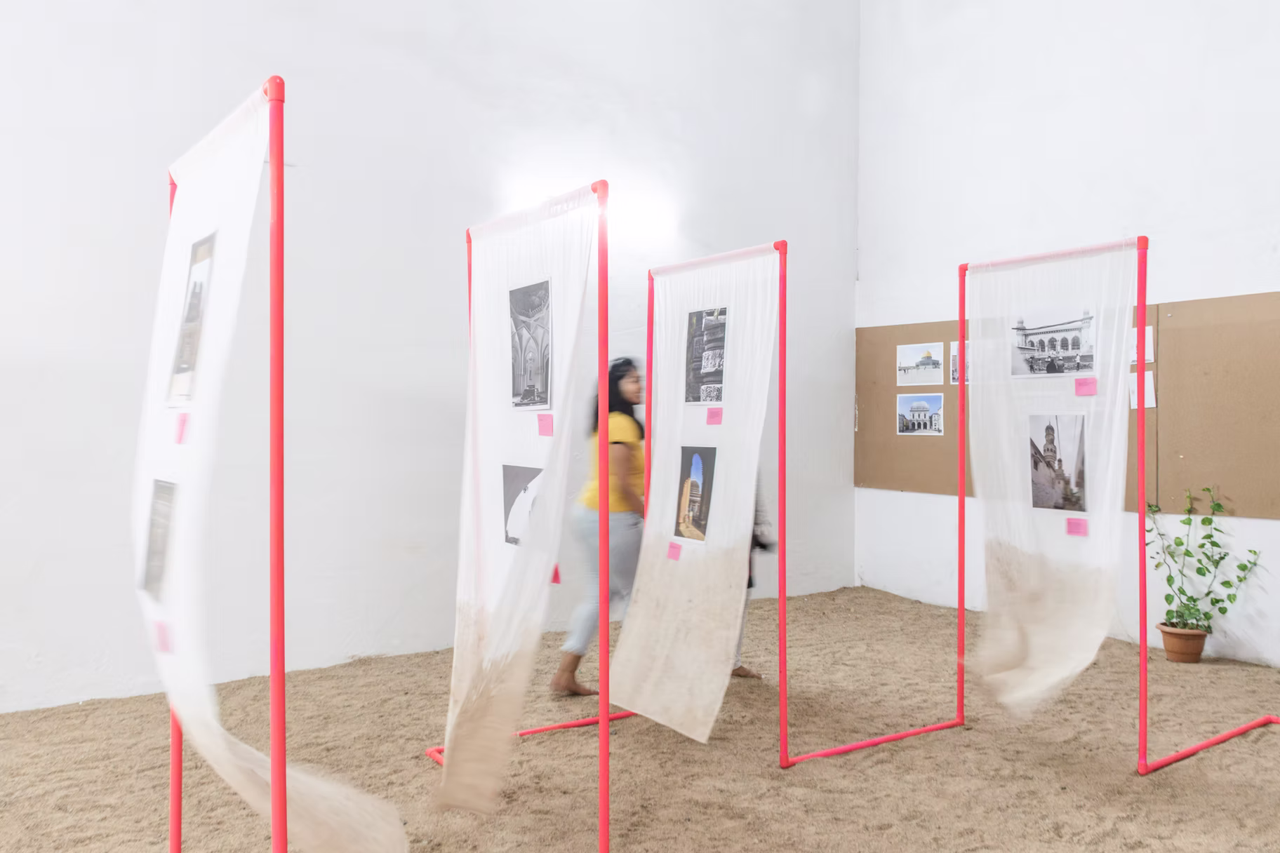 Visitor walking past sheer panels on pink frames, prints fluttering above sand with a potted plant nearby, Hyderabad gallery