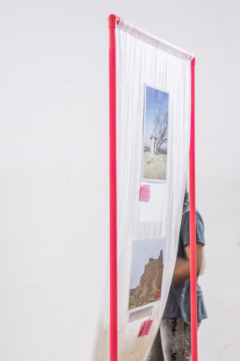 Single red-framed gauze panel with two photo prints and a pink tab, a person half-hidden behind it, white Hyderabad gallery