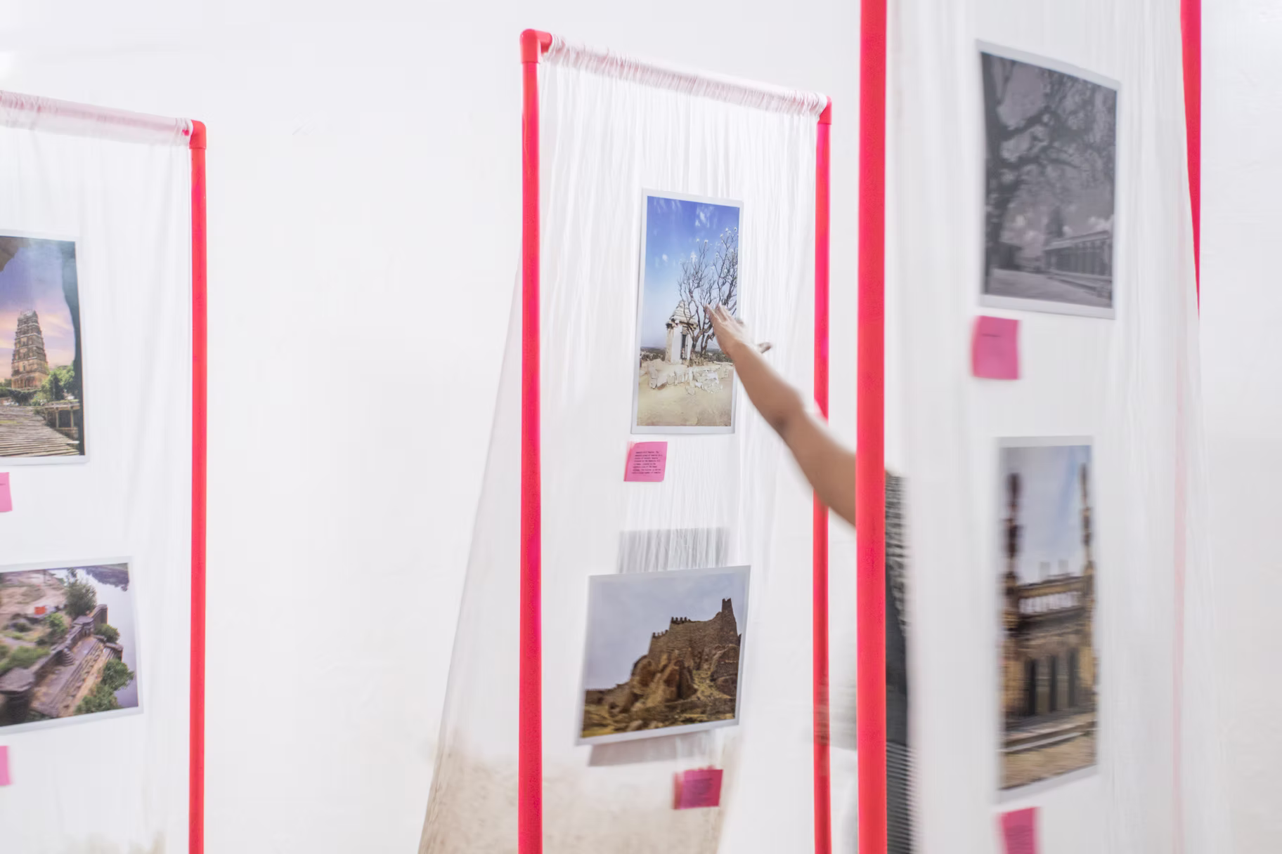 Heritage-monument photo prints pinned to white gauze panels with pink tabs, a hand reaching toward one, Hyderabad gallery