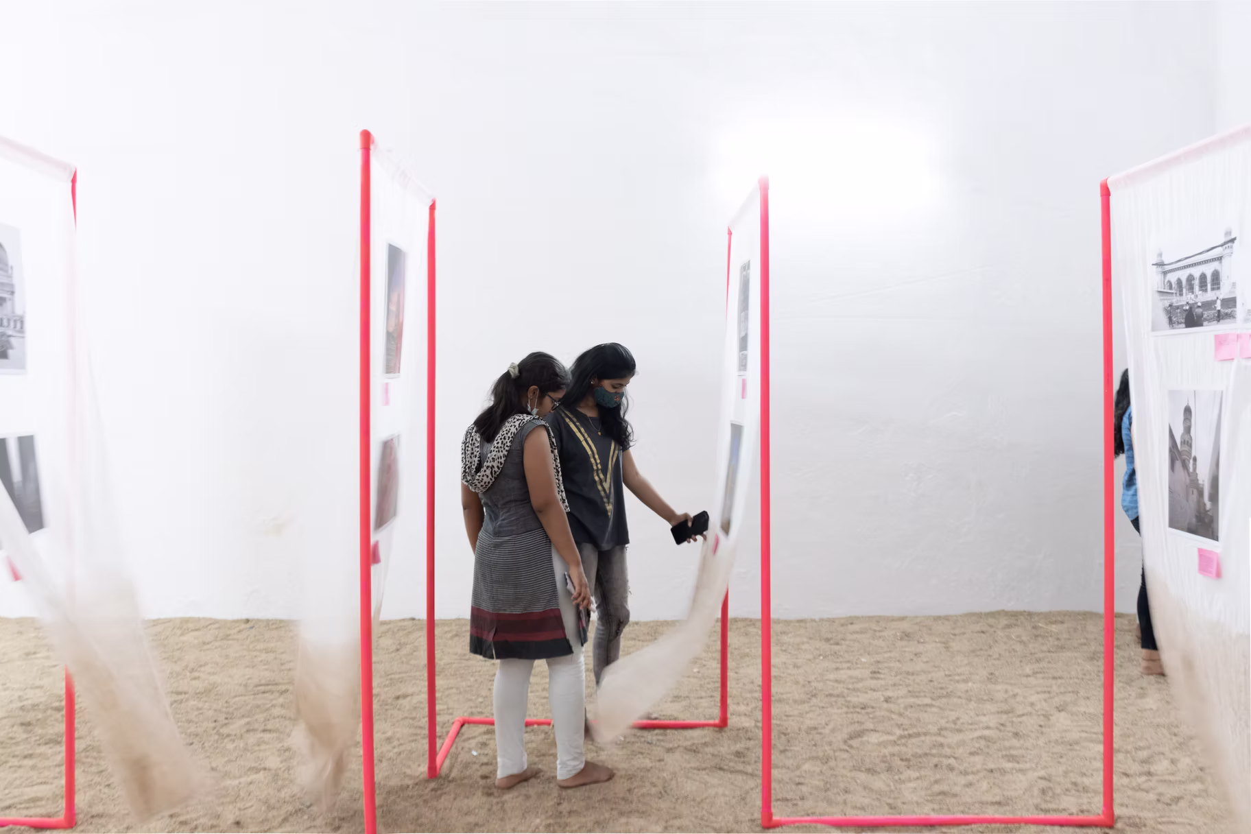 Two visitors view a photo installation of red-bordered translucent panels and trailing fabric on a sand-covered floor