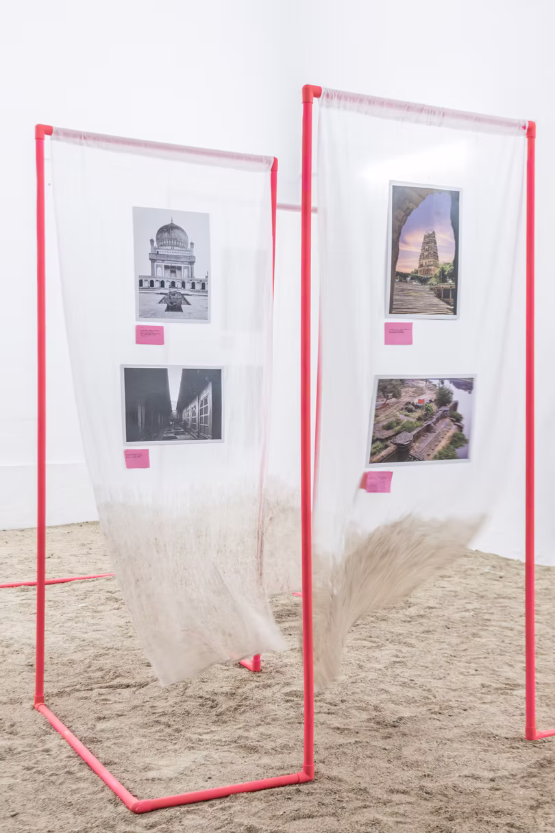 Pair of red-framed gauze panels pinned with temple and ruin prints, fabric spreading across sand in a Hyderabad gallery
