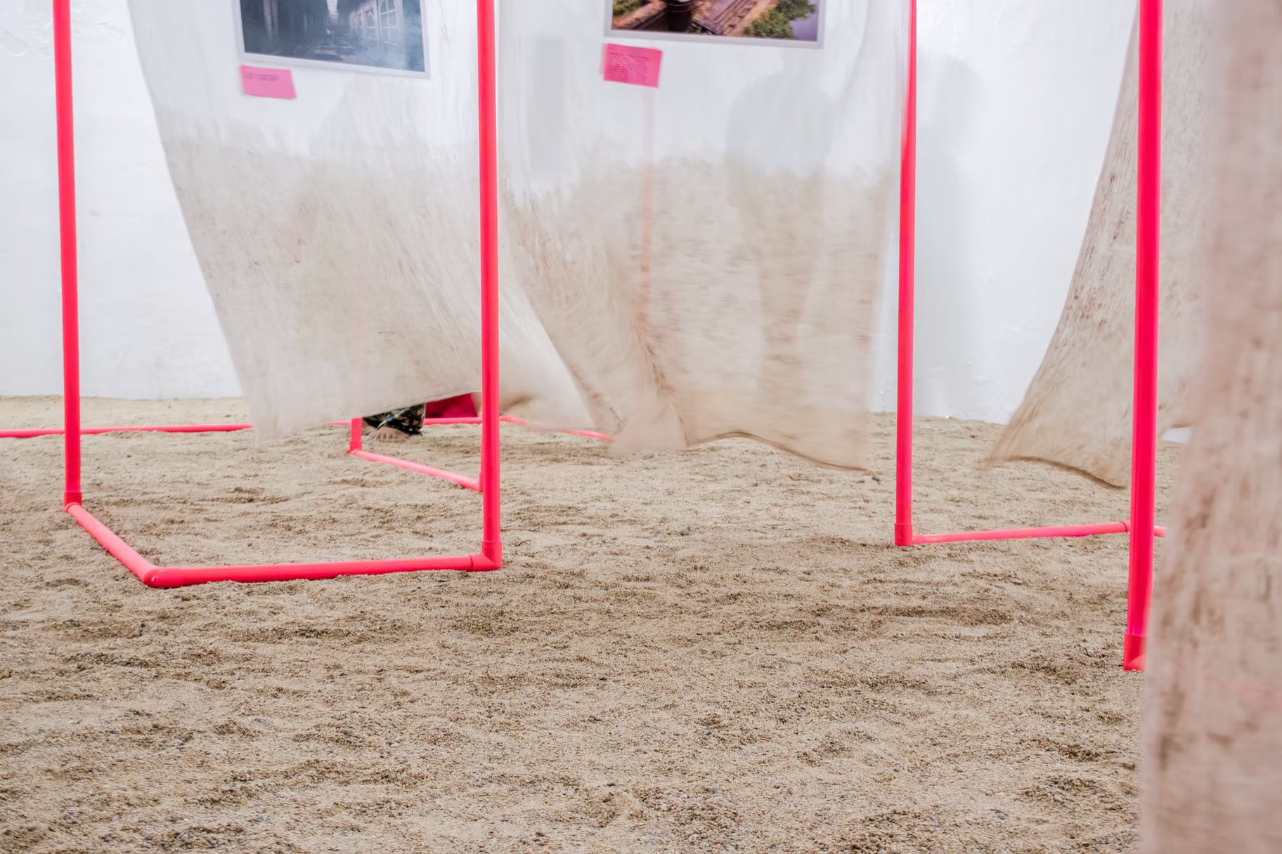 Low view of hot-pink pipe frame bases and gauze hems meeting a sand-covered gallery floor, Hyderabad installation detail