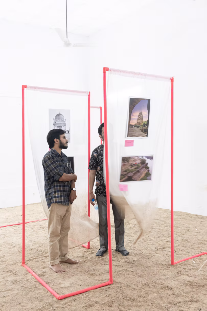Two men standing among red doorway frames hung with photographs over sand in a white-walled Hyderabad exhibition