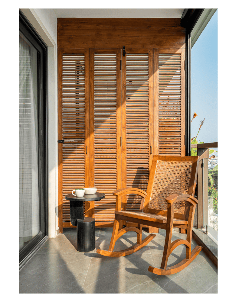 Sunlit balcony nook with wooden louvred screen, a cane-back rocking chair and a black drum side table set for coffee