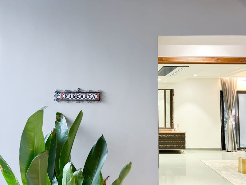 Carved metal nameplate reading NISCHITA mounted on a grey wall beside a leafy plant near a bright entryway, Hyderabad home
