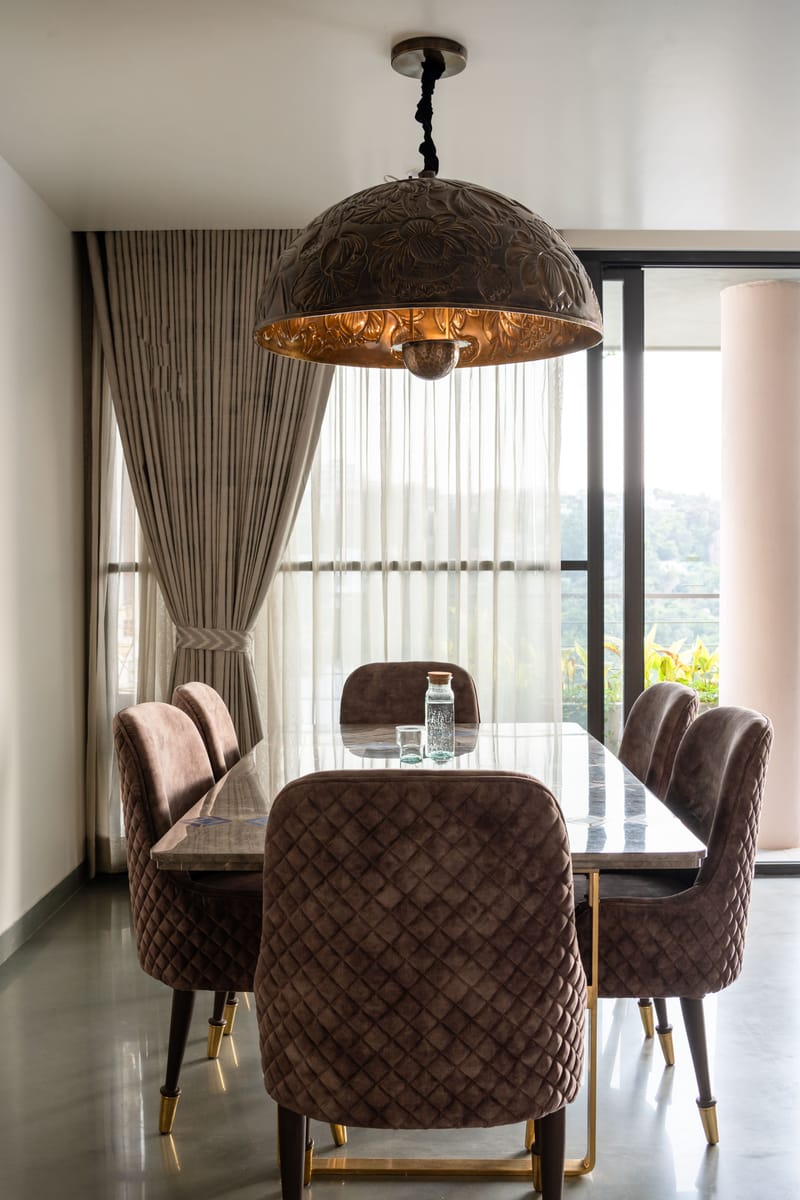 Six-seater dining set, Hyderabad — quilted brown velvet chairs on brass-tipped legs under a domed copper pendant by sheer-draped windows