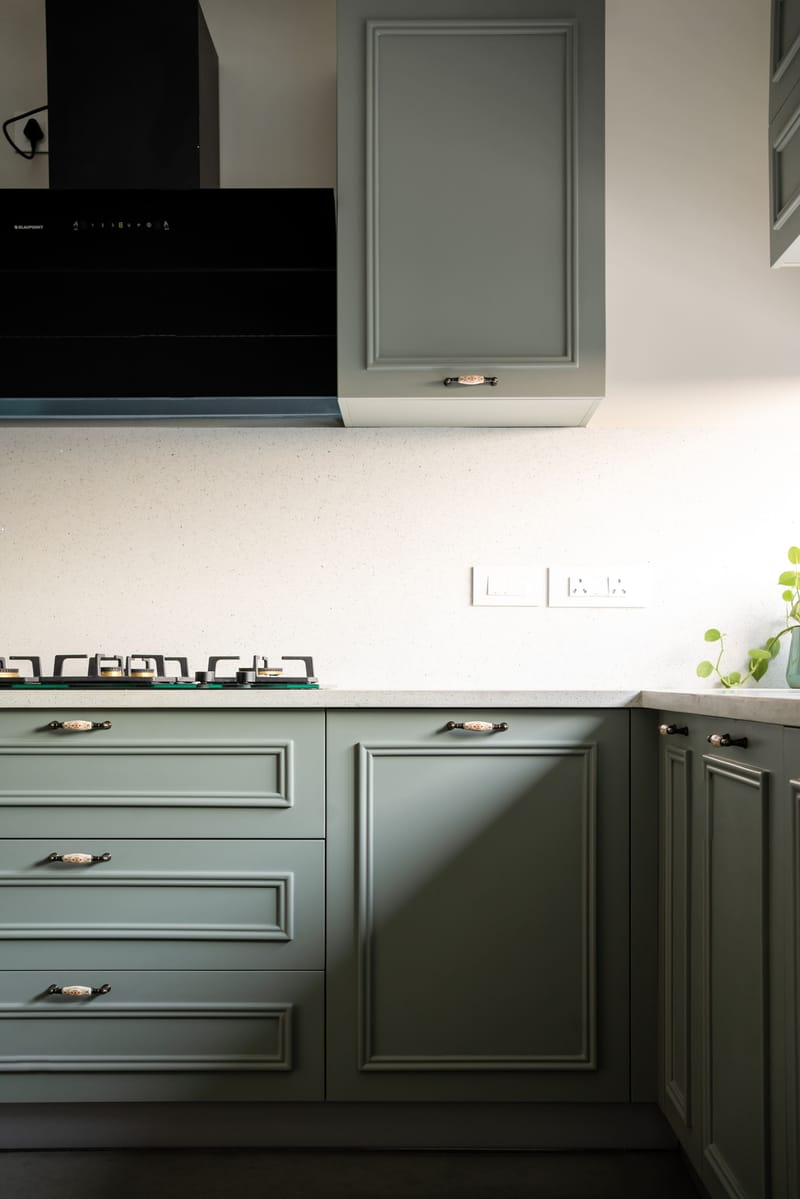 Sage-green kitchen detail, Hyderabad — paneled cabinets with cup pulls, black wall chimney and gas hob on a speckled quartz top