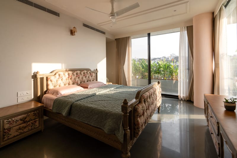 Spacious master bedroom, Hyderabad — tufted floral carved bed, green bedspread and full-height windows to a leafy balcony