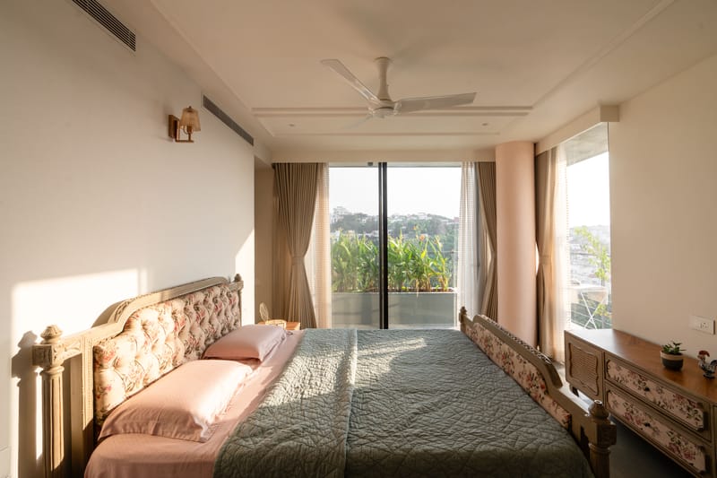 Master bedroom in Hyderabad with a carved wood bed, sage-green quilt and balcony doors framing a hillside garden view