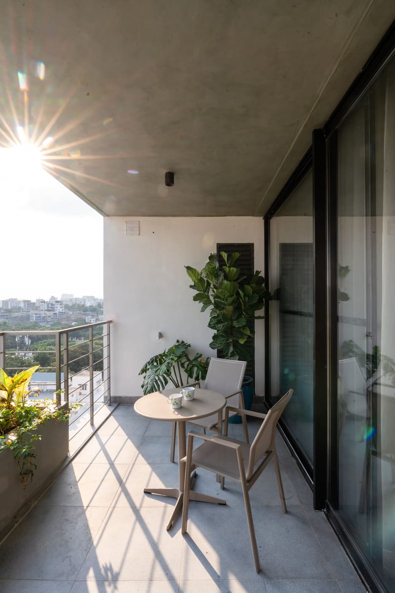 Sunlit balcony with two cane-style chairs, round table and tall indoor plant framed by glass sliding doors, Hyderabad