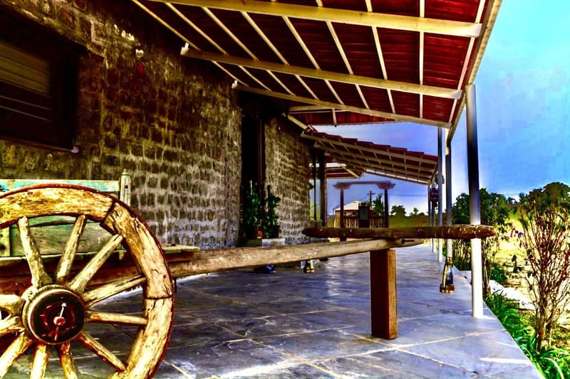 Farmhouse stone verandah in Hyderabad with sloped timber roof and an antique wooden bullock-cart wheel on slate flooring