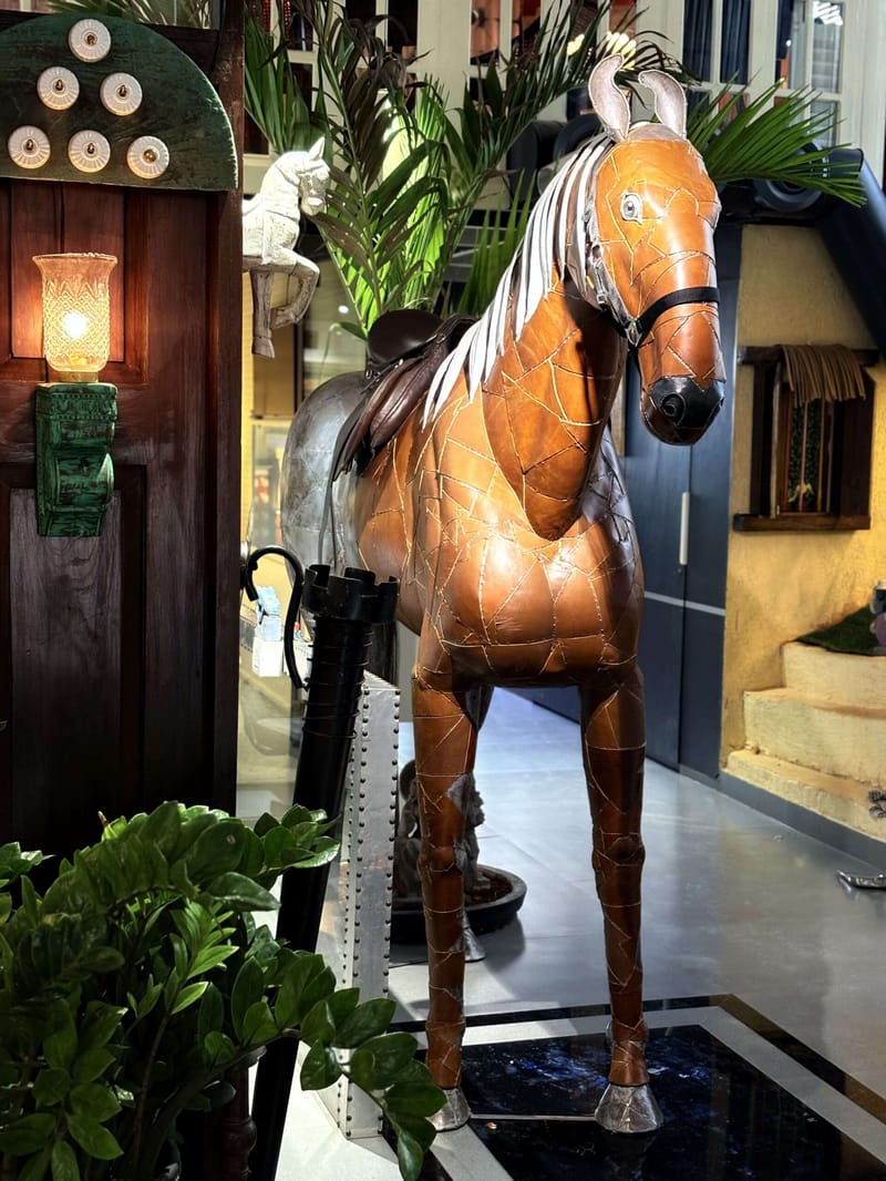 Life-size leather-clad horse sculpture displayed among potted palms and a green wall lantern in a decor showroom