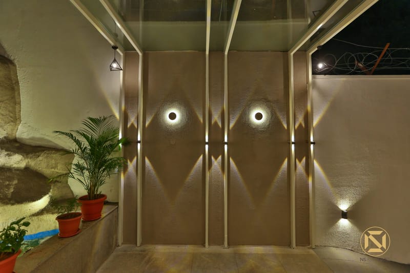 Textured taupe feature wall with recessed circular wall lights and potted palms in a Hyderabad home entryway courtyard