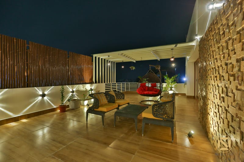Rooftop terrace lounge at night with hanging swing chair, cane seating and a textured stone wall on a wood deck, Hyderabad