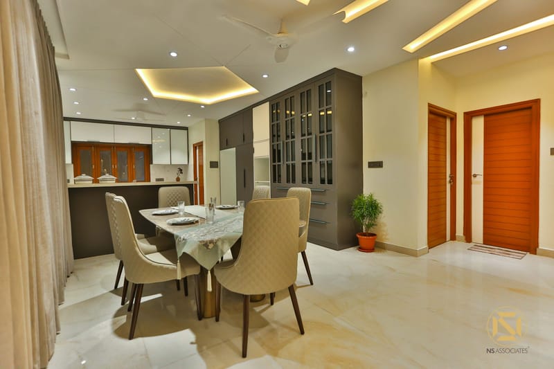 Dining area with beige tufted chairs, glass-front crockery cabinets and modular kitchen behind warm wooden doors