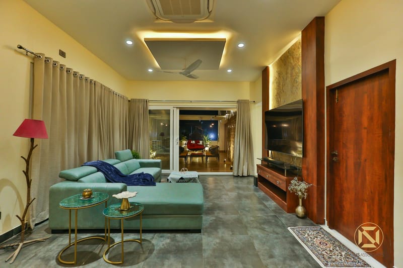 Living room with teal sectional sofa, red floor lamp, wood TV unit and grey tile floor opening to a lit terrace, Hyderabad