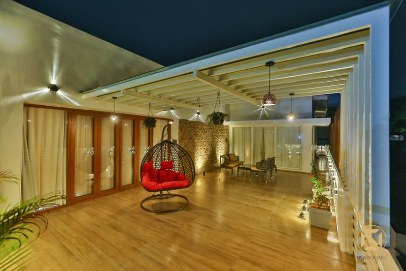 Rooftop terrace under a white pergola with a red-cushioned egg swing and stone wall over a wooden deck at night, Hyderabad