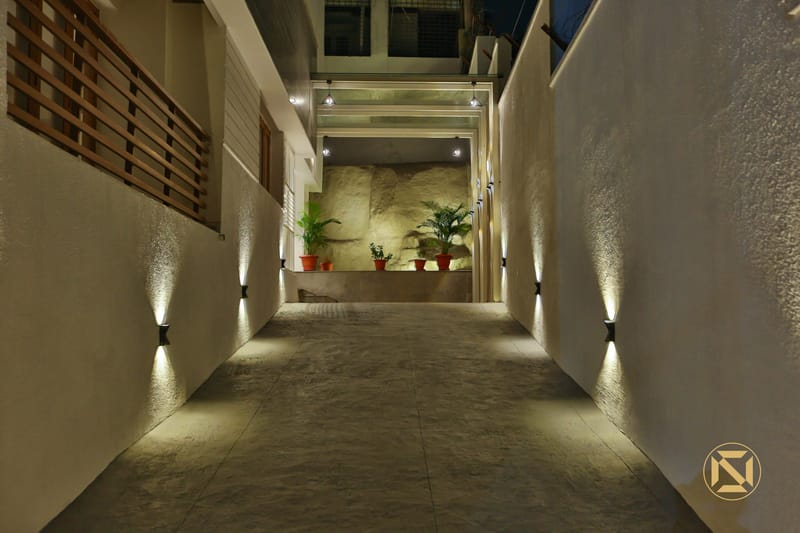 Outdoor entry passage at night lit by up-and-down wall lights, with potted plants against a rough stone niche, Hyderabad