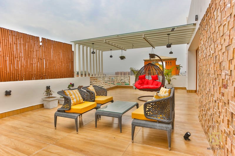 Open terrace seating with woven rattan armchairs, yellow cushions and a red hanging swing by a brick wall, Hyderabad