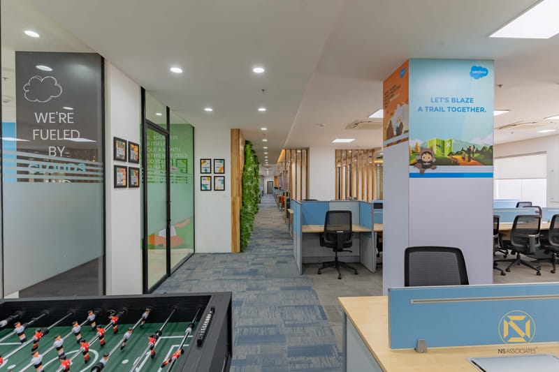 Office breakout zone with foosball table, framed art, glass cabins and blue reception pod under recessed lights, Hyderabad