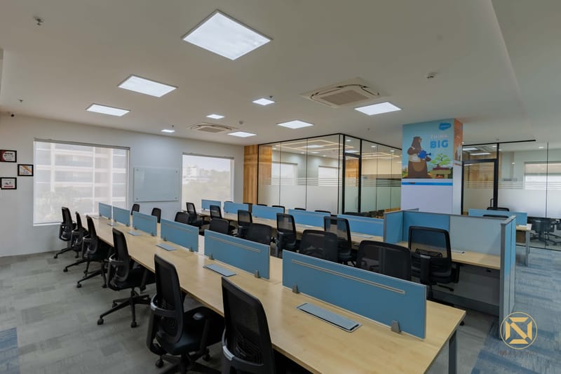 Long rows of office desks under recessed ceiling light panels, blue dividers and a glass meeting cabin, Hyderabad