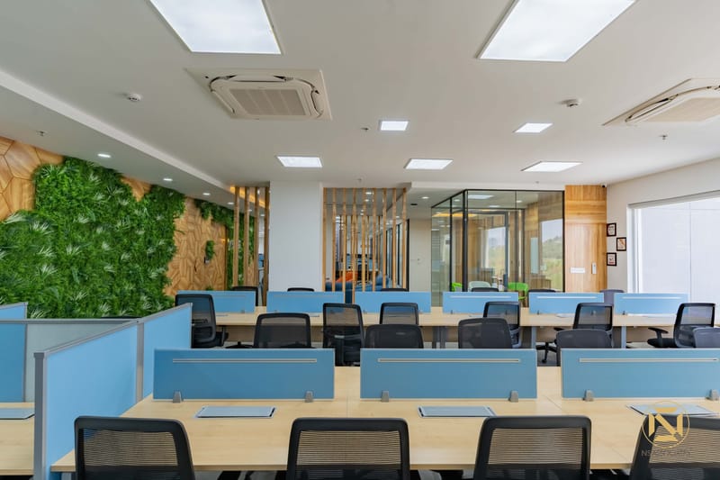 Open-plan office workstations with blue desk dividers, mesh chairs and a green moss wall on wood panelling, Hyderabad