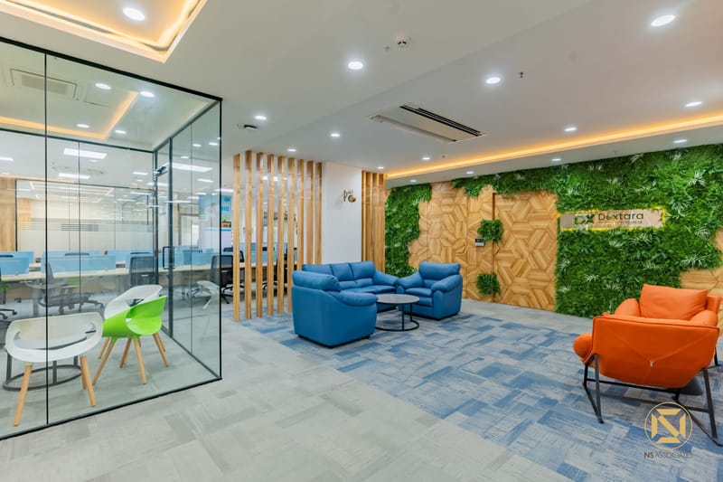 Breakout lounge with blue sofa, orange chair, vertical green wall and a glass meeting cabin with wood-slat screen, Hyderabad
